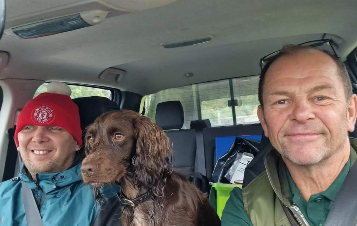 Volunteer Paul, senior ranger Dave and his dog sit in the front of a car smiling at the camera